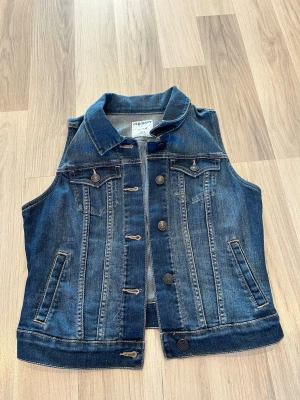Mörkblå ärmlös jeansväst  - Mörkblå ärmlös jeansjacka med klassisk krage, knappar framtill och två bröstfickor med lock. Snygga sömmdetaljer och två sidofickor. Perfekt att bära över en hoodie eller t-shirt för en avslappnad stil.