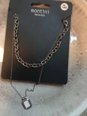 Silverfärgat halsband från Montini - Snyggt halsband i silverfärg från Montini med en kombination av grov kedja och tunnare kedja. Halsbandet har en rektangulär vit detalj som hänge, vilket ger en modern och trendig look. Perfekt accessoar för att lyfta din outfit.