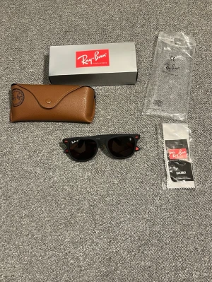  Ray-Ban  solglasögon - Klassiska svarta Ray-Ban Wayfarer solglasögon med mörka glas och röda detaljer på bågarna. Säljs med originalfodral, putsduk och box. Perfekt accessoar för dig som vill ha en tidlös och ikonisk look.