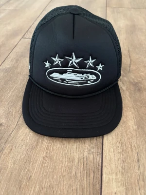 Corteiz (CRTZ) Trucker Cap – Svart – Nyskick - Passa på att lägga vantarna på en klassisk trucker-keps från Corteiz! En riktig streetwear-staple med den ikoniska stjärn-logotypen och detaljerna som syns på bild. Kepsen är i en stilren svart färg med den klassiska "rope"-detaljen vid brättet. Den är perfekt för att komplettera din outfit och ger direkt den rätta streetwear-looken.  Märke: Corteiz (CRTZ)  Modell: Trucker Cap  Skick: [Ange skick, t.ex. Nyskick/Använd ett fåtal gånger]  Färg: Svart 