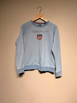 Ljusblå sweatshirt från GANT - Ljusblå sweatshirt från GANT med broderad logga och sköld med stjärnor och ränder på bröstet. Tröjan har rund halsringning, ribbade muddar och avslappnad passform. Perfekt för en casual och sportig stil.