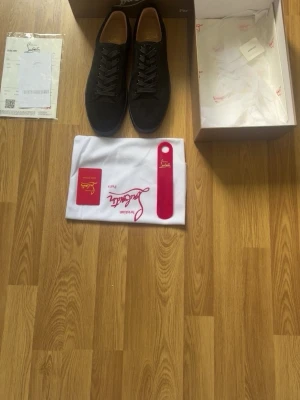 Svarta sneakers från Christian Louboutin - Stilrena svarta sneakers från Christian Louboutin med klassisk röd sula och svarta skosnören. Skorna har låg profil och är tillverkade i mocka med läderdetaljer. Medföljer originalkartong, dustbag och skohorn med Louboutin-logga.