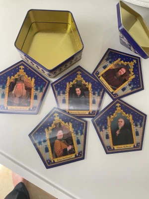 Harry Potter Chocolate Frog ask & kort - Säljer en ikonisk Harry Potter Chocolate Frog plåtburk i blått och guld, tillsammans med fem samlarkort med karaktärer från serien. Perfekt för fans eller samlare som vill ha en snygg pryl till hyllan eller förvaring.