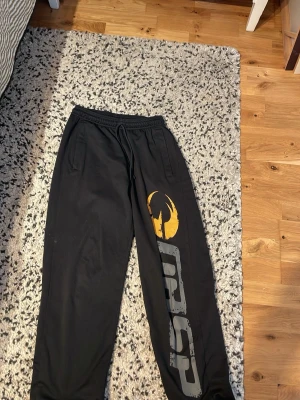 Svarta mjukisbyxor från GASP - Svarta mjukisbyxor från GASP med resår och dragsko i midjan. Stora tryck i grått och gult längs ena benet och två sidofickor. Perfekta för chill eller gym. Materialet känns mjukt och bekvämt.