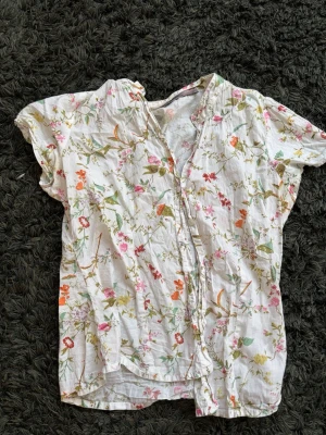 Vit blommig kortärmad blus - Söt vit blus med färgglatt blommönster i rosa, orange och grönt. Blusen har korta ärmar och knäppning framtill 