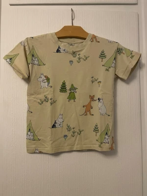 Gul Mumin t-shirt med tryck - Söt gul t-shirt med korta ärmar och färgglatt Mumin-tryck över hela plagget. Motiven inkluderar Mumin, Snusmumriken och andra karaktärer bland tält och träd. Perfekt för dig som gillar Mumin och vill ha en lekfull stil.