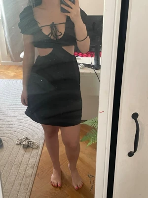 Svart kort klänning med cut-outs - Snygg svart kort klänning med trendiga cut-outs i midjan och snörning framtill. Klänningen har puffiga korta ärmar och en figurnära passform som ger en modern look. Perfekt för dig som vill sticka ut med en unik och stilren outfit.