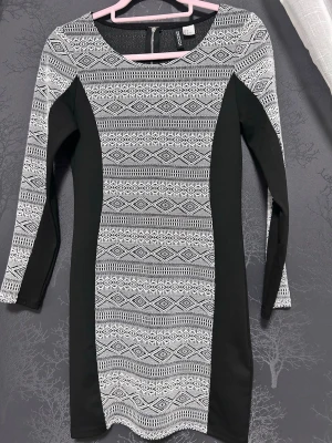 Svartvit mönstrad klänning H&M - Snygg svartvit kortklänning från H&M med grafiskt mönster och svarta paneler i sidan. Klänningen har lång ärm, rund halsringning och dragkedja bak. Perfekt för dig som gillar stilrena kontraster och en figurnära passform.