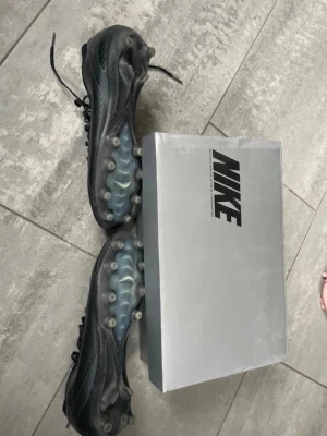 Svarta Nike fotbollsskor med dobbar - Säljer ett par svarta Nike fotbollsskor med snörning och låg profil. Skorna har en modern design med strukturmönster på ovandelen och fasta dobbar för bra grepp på planen. Perfekta för dig som vill ha stilrena och funktionella skor till match eller träning. Elite, samma modell som proffs använder (den mest exklusiva modellen som Nike har). Originalkartong ingår. Skopåsen medföljer men har en reva, se bild.