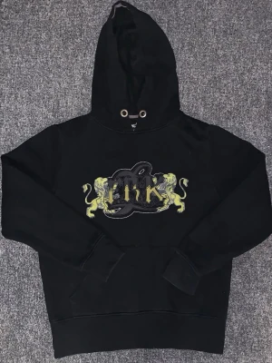 Svart hoodie från Identify med tryck - Svart hoodie från Identify med stort tryck på bröstet i gult och grått, två lejon och texten 'Link'. Hoodien har huva med metallöljetter och ribbade muddar vid ärmar och nederkant. Perfekt för en streetwear-look.