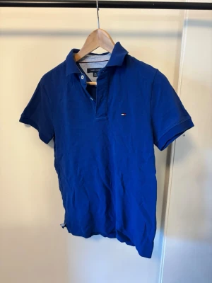 Blå pikétröja från Tommy Hilfiger - Snygg blå pikétröja från Tommy Hilfiger med klassisk krage och korta ärmar. Tröjan har en liten logga på bröstet och är gjord i mjuk bomull. Perfekt till jeans eller shorts för en avslappnad look.