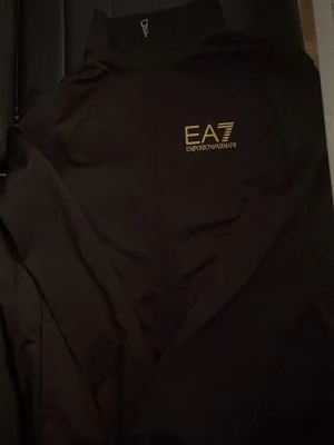 Svart ziptröja från Emporio Armani - Snygg svart ziptröja från Emporio Armani EA7 med guldfärgad logga på bröstet. Tröjan har hög krage och dragkedja framtill. Perfekt för en sportig och stilren look.