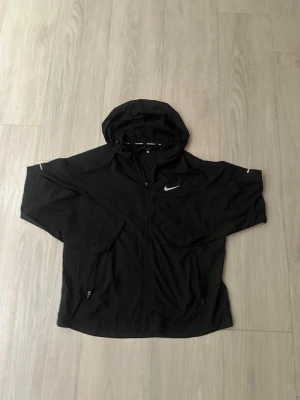 Svart vindjacka från Nike med huva - Snygg svart vindjacka från Nike med dragkedja framtill och huva. Jackan har två fickor med dragkedja och reflekterande detaljer på ärmarna samt Nike-logga på bröstet. Tillverkad i lätt och tunt material som passar perfekt för blåsiga dagar.