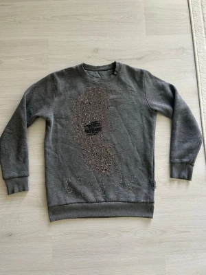 Grå sweatshirt med döskalle från Philipp Plein - Cool grå sweatshirt från Philipp Plein med en stor döskalle framtill i glittriga nitar och texten 'PHILIPP PLEIN' under motivet. Tröjan har rund halsringning och långa ärmar. Perfekt för dig som gillar edgy streetstyle.