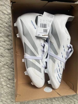 Adidas Predator vita fotbollsskor - Snygga vita Adidas Predator fotbollsskor med silvergrå detaljer och blå accenter. Skorna har klassisk snörning och är tillverkade i syntetmaterial med mönstrad ovandel för extra grepp. Perfekta för dig som vill sticka ut på planen.