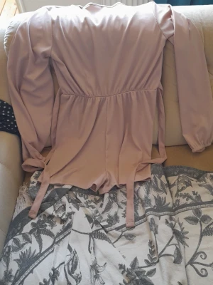 Ljusrosa långärmad jumpsuit - Supersnygg ljusrosa jumpsuit med långa ärmar och knytband i midjan. Kort modell med resår i midjan för en skön passform. Tillverkad i ett mjukt och stretchigt tyg som känns bekvämt hela dagen.