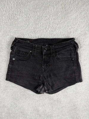 True Religion Jennie Curvy Mid Rise Skinny Jeansshorts storlek 25 Svarta - Snygga svarta denimshorts med klassisk femficksdesign och knappgylf. Shortsen har bälteshällor och är korta i modellen, perfekta för varma dagar. Passar dig som gillar en enkel och stilren look.