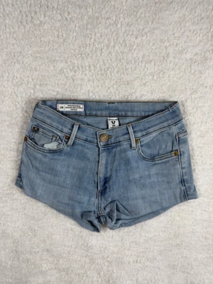 True Religion Liv Low Rise Relaxed Skinny Shorts W25 - Säljer ett par ljusblå denimshorts från True Religion med låg midja och klassisk femficksdesign. Shortsen har knappgylf och bälteshällor, perfekta för en avslappnad och trendig look.
