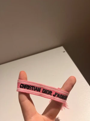 Christian Dior rosa tygarmband - Säljer ett rosa tygarmband från Christian Dior med texten 'Christian Dior J'Adior' i svart. Armbandet har en vävd struktur och avslutas med tofsar i båda ändar. Perfekt accessoar för dig som gillar exklusiva och trendiga detaljer.