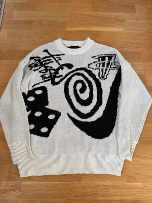 Vit och svart stickad tröja Stüssy - Säljer en stickad tröja från Stüssy i vitt med svart grafiskt mönster. Tröjan har rund hals, ribbade muddar och en oversized passform. Perfekt för dig som gillar streetwear och unika prints.