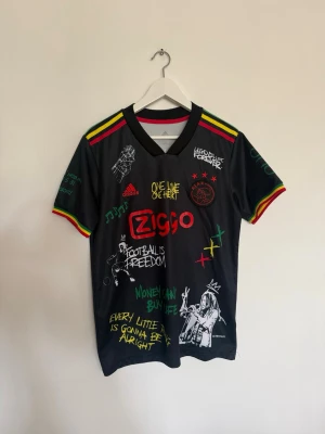 Svart Ajax x Bob Marley tröja - Unik svart Ajax x Bob Marley matchtröja från Adidas med färgglada detaljer i rött, gult och grönt. Tryck med Ziggo-logga, Bob Marley-motiv och texter som 'Football is Freedom' och 'Live the life you love'. Korta ärmar och V-ringning. Perfekt för fotbollsfans och streetwear.