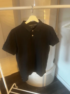 Svart pikétröja från Sail Racing - Klassisk svart pikétröja från Sail Racing med krage och två knappar framtill. Tillverkad i bomull för en skön och avslappnad känsla. Perfekt till jeans eller shorts för en clean look.