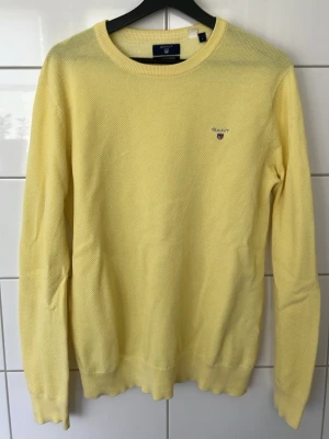 Gul stickad tröja från Gant - Ljusgul stickad tröja från Gant med rund halsringning och diskret logga broderad på bröstet. Tröjan har långa ärmar och ribbade muddar vid ärmslut och nederkant. Perfekt för en fräsch och stilren look.
