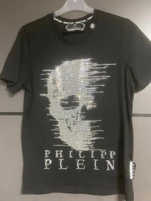 Svart t-shirt med döskalle från Philipp Plein - Snygg svart t-shirt från Philipp Plein med glittrande döskallemotiv framtill och texten 'PHILIPP PLEIN' i stenar. Klassisk rund halsringning och korta ärmar. Perfekt för dig som gillar statement-plagg och exklusiv design.