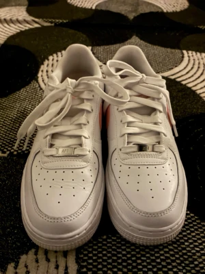Vita sneakers med rosa swoosh - Fräscha vita Nike sneakers med klassisk rosa swoosh på sidan. Skorna har lågt skaft, perforerad tåbox och vita skosnören. Tillverkade i läder med detaljer i gummi och textil. Perfekt för dig som gillar en clean och sportig look med en touch av färg.