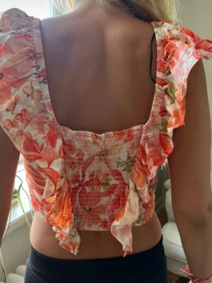 Blommig volangtopp med rysch - Supersöt blommig topp med volanger och ryschdetaljer. Toppen har en croppad passform, breda axelband och är sydd i ett tunt, lätt material med orange, röda och gröna toner. Perfekt för sommarens alla tillfällen!