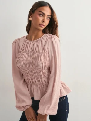 Ljusrosa smockad blus med volang från nelly - Supersöt ljusrosa blus med smockad midja och volang nertill. Blusen har långa ärmar och rund halsringning. Materialet ser ut att vara mjukt och lite glansigt, vilket ger en fin känsla. Perfekt för dig som gillar romantisk och feminin stil.