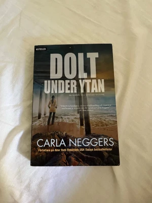 Dolt under ytan av Carla Neggers - Spännande deckare av Carla Neggers med omslag i mörka toner och motiv av strand och hav. Pocketbok med tydlig titel i vitt och författarens namn i guld. Perfekt för dig som gillar mysterier och thrillers.