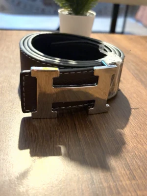 Bälte  - Snyggt brunt bälte i skinn med ett stort, silverfärgat H-format spänne. Bältet har vita sömmar längs kanterna som ger en clean kontrast. Perfekt accessoar för att lyfta din outfit.