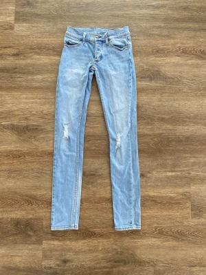 Cheap Monday jeans - stl 24/32 - Säljer ett par ljusblå skinny jeans med slitningar på knäna och unika tryckknappar med döskalle och skelettmotiv. Jeansen har klassisk femficksdesign och orangea sömmar. Perfekta för dig som gillar detaljer och streetstil.