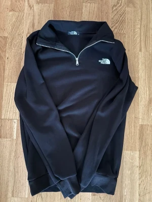 Svart half-zip tröja The North Face - Svart half-zip tröja från The North Face med hög krage och dragkedja framtill. Tröjan har en stilren design med logga på bröstet och är tillverkad i ett mjukt material. Perfekt för lager-på-lager och enkel att matcha med andra plagg. Pris kan diskuteras vid snabb affär! 
