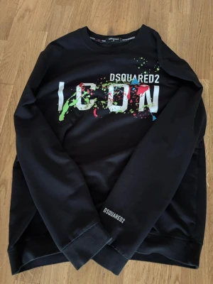 Svart ICON hoodie från Dsquared2 - Säljer en svart hoodie från Dsquared2 med stor 'ICON'-logga och färgstänk i grönt, rött och blått på bröstet. Tröjan har rund halsringning och tryckt Dsquared2-detalj på ärmen. Perfekt för dig som gillar streetwear och vill sticka ut. Hej bli inte lurade det står xl på taggen men det är M! Pris kan diskuteras vid snabb affär 