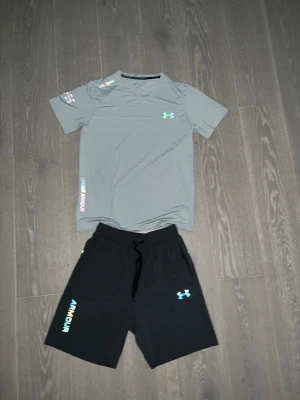 Under Armour träningsset t-shirt & shorts - Snyggt träningsset från Under Armour med en ljusgrå t-shirt och svarta shorts. T-shirten har logga på bröstet och tryck på ärmen, medan shortsen har dragsko i midjan, logga och texten 'ARMOUR' på benet. Perfekt för gymmet eller löpning.