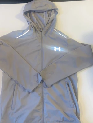 Ljusgrå vindjacka från Under Armour - Snygg ljusgrå vindjacka från Under Armour med huva och dragkedja framtill. Jackan har två sidofickor och är tillverkad i ett lätt, vindtätt material som passar perfekt för blåsiga dagar. Stort logotryck på bröstet ger en sportig look.storlek S/M