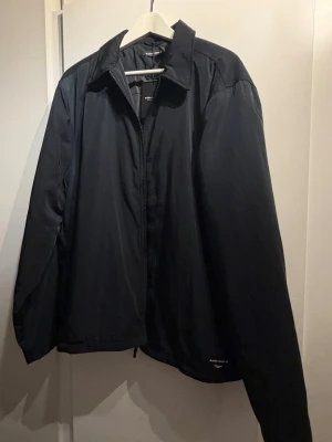Svart vindjacka med krage - Stilren svart vindjacka med klassisk krage och dragkedja framtill. Jackan har en enkel design utan synliga loggor och är tillverkad i ett lätt, vindtätt material. Perfekt för blåsiga dagar och enkel att matcha med olika outfits. Nypris-1349 