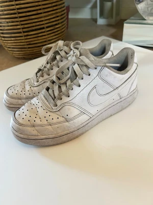 Vita Nike sneakers med grå detaljer - Säljer ett par klassiska vita Nike sneakers med grå detaljer och snörning. Skorna har perforerad tåbox för ventilation och en låg siluett som passar till många olika stilar. Tillverkade i läder med textilfoder och gummisula.