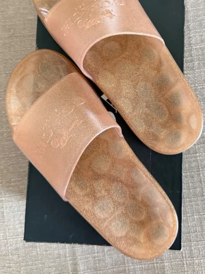 Glittriga  slides från Coach - Säljer ett par glittriga beige sandaler från Coach. De har en transparent finish med skimrande effekt och Coach-logga på ovandelen. Tillverkade i syntetmaterial med Coachs signaturmönster på sulan. Perfekta för en trendig och avslappnad look. OBS! hård material 