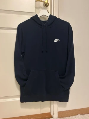 Mörkblå hoodie från Nike - Snygg mörkblå hoodie från Nike med klassisk logga i vitt på bröstet. Hoodien har huva med dragsko och en stor magficka framtill. Tillverkad i mjukt material som känns skönt mot huden. Perfekt för en avslappnad och sportig stil. Storlek S