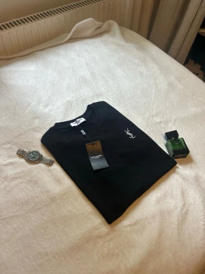Svart YSL t-shirt med logga - Snygg svart t-shirt från Yves Saint Laurent med vit YSL-logga broderad på bröstet. Klassisk rund hals och korta ärmar. Tillverkad i mjuk bomull som känns skön mot huden. Perfekt för dig som vill ha en stilren och trendig look.