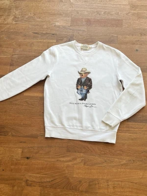 Vit sweatshirt Polo Ralph Lauren - Vit sweatshirt från Polo Ralph Lauren med tryck av en björn i cowboy-outfit på bröstet. Klassisk rund halsringning och ribbade muddar vid ärmslut och nederkant. Tillverkad i mjukt bomullsmaterial för en skön och avslappnad look.