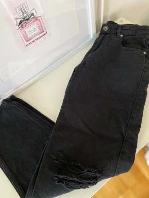svarta raka jeans med slitningar  - Säljer ett par svarta jeans med hög midja och slitningar vid knät. Jeansen har klassisk femficksmodell, bälteshällor och knappgylf. Materialet är denim med stretch för skön passform. Perfekta för en trendig och avslappnad look.
