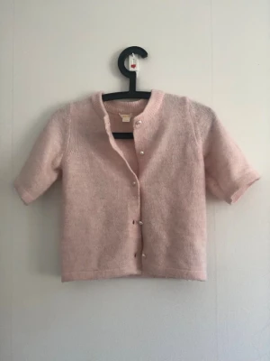 Ljusrosa kofta med kort ärm - Söt ljusrosa kofta med korta ärmar och rund hals. Koftan har pärlknappar framtill och är stickad i ett mjukt material.💗