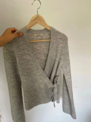 Grå omlottkofta med knytband - Mysig grå omlottkofta i mjukt stickat material. Koftan har långa ärmar och knyts i sidan med ett brett band för en snygg och avslappnad look. Perfekt att slänga över en topp för extra lager och stil.