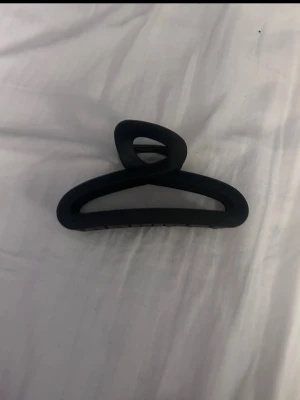 Svart hårklämma i matt plast - Säljer en stilren svart hårklämma i matt plast. Klämman har en bred, avlång form med öppna sidor och stark fjäder, perfekt för att hålla upp håret på ett snyggt sätt. Enkel att matcha med olika outfits och passar till alla hårtyper.