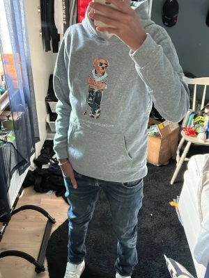 Grå hoodie Polo Bear Ralph Lauren - Snygg grå hoodie från Ralph Lauren med Polo Bear-tryck på bröstet. Hoodien har huva och en stor magficka framtill. Tillverkad i mjukt material och har ribbade muddar vid ärmar och nederkant. Perfekt för en chill och avslappnad stil.