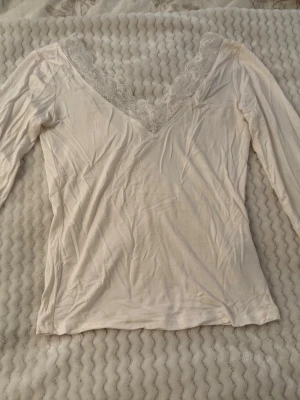 Vit långärmad topp med spets - Säljer en vit långärmad topp med djup v-ringning och fina spetsdetaljer runt halsen. Toppen är mjuk och har en feminin look som passar perfekt till jeans eller kjol. Enkel men ändå elegant design. Använd 2 gånger behöver strykas bara annars som ny 